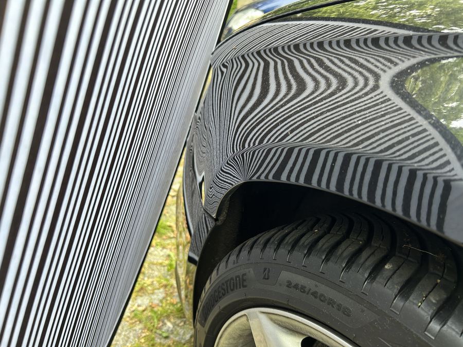 Vorderer Kotflügel eines schwarzen Audi A4 am Radlauf unter der Dellenleuchte, die Streifen laufen an der eingedrückten Stelle in Wellen auseinander