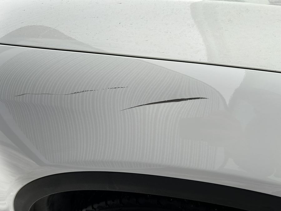 Vorderer Kotflügel eines weißen Mercedes über dem Radlauf mit zwei langen Kratzern im Lack, in der Spiegelung einer Streifenleuchte zeichnet sich eine leichte Verformung ab