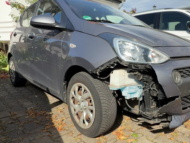 Vordere rechte Ecke eines grauen Hyundai i10 mit abgerissener Stoßfängerabdeckung, freiliegendem Wischwasserbehälter und beschädigtem Scheinwerfer, Reparaturkosten laut Gutachten 6.620 Euro brutto